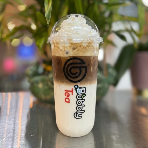Layered Bubbly Tea drink with whipped cream on top, against a blurred green plant background.