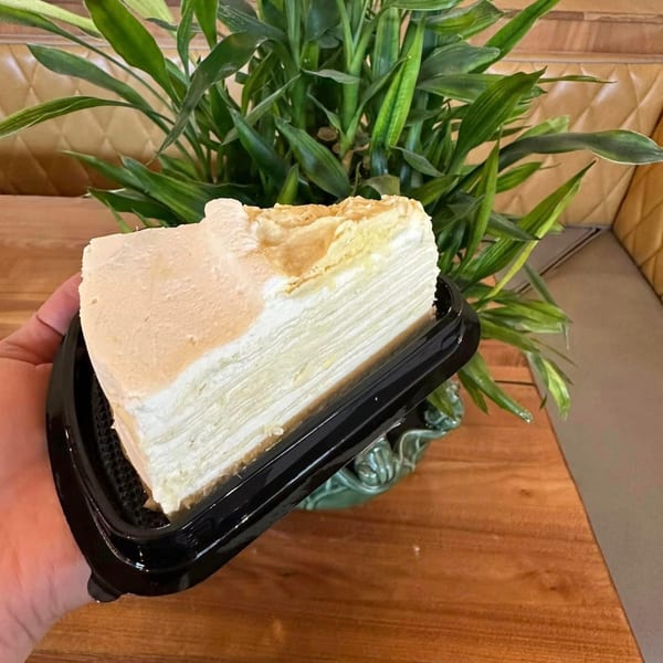 A hand holds a slice of layered crepe cake with white cream, against a plant and wooden table.