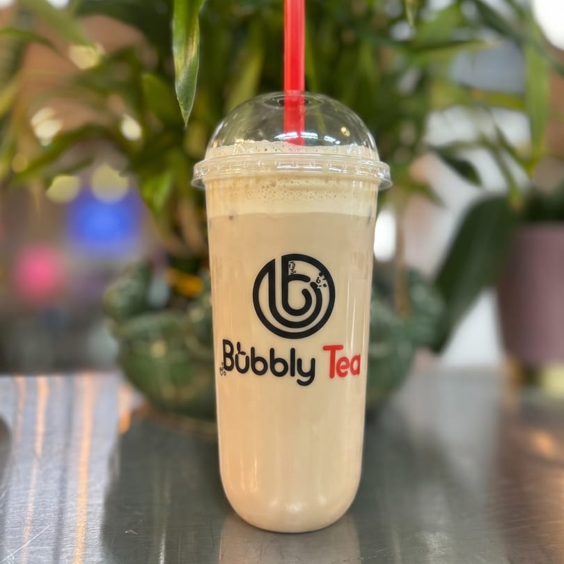 A Bubbly Tea branded cup of light brown bubble tea with a red straw on a reflective surface.