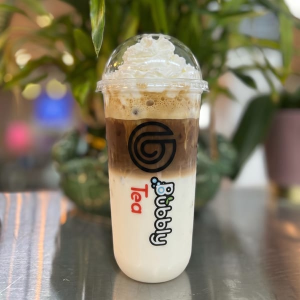 Bubbly Tea's layered iced coffee with whipped cream in a clear plastic cup on a metallic surface.